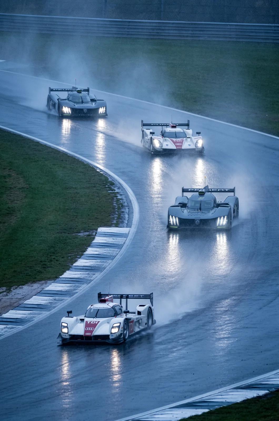 Four prototype race cars kick up spray on a wet, curving track, headlights glowing in the blue dusk.